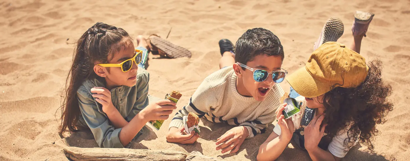 Three laughing kids laying on sand eating Nature Valley granola bars.