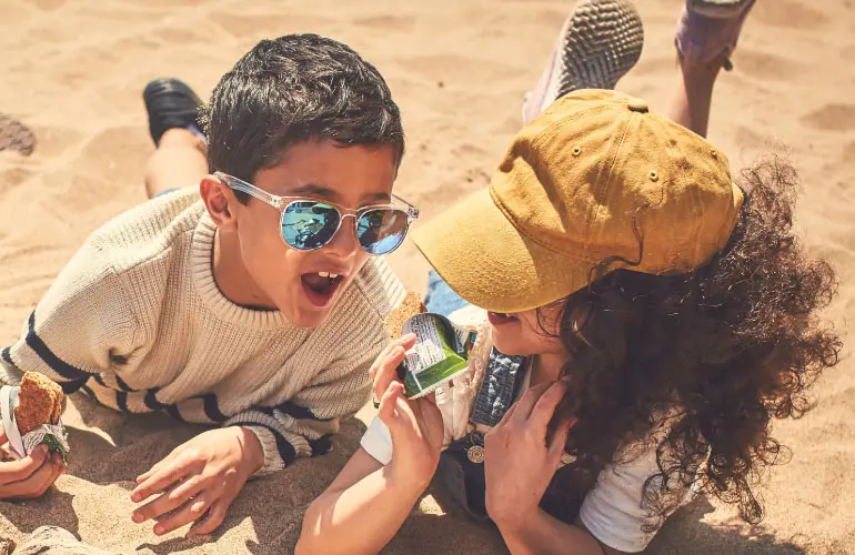 Three laughing kids laying on sand eating Nature Valley granola bars.