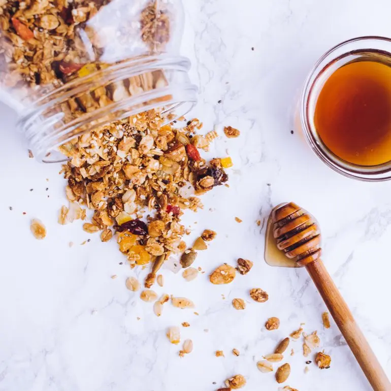 Granola spills out of a glass jar onto a marble countertop. A bowl of honey sits nearby.