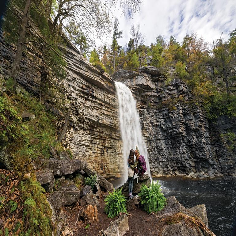 Two people stand close together while looking towards a waterfall.