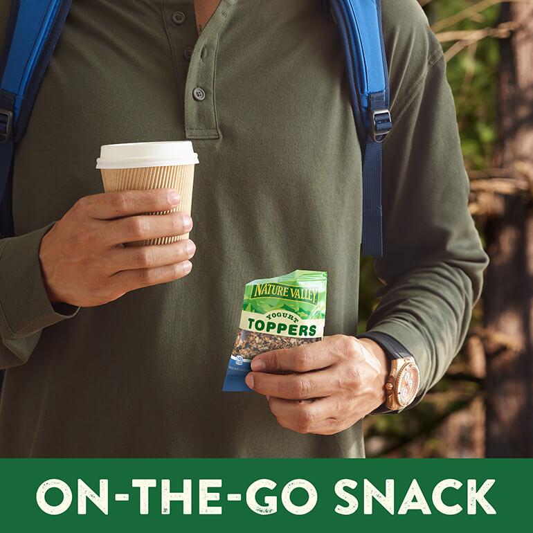 A close up of a man in a green top holding a coffee cup and a pouch of Nature Valley Yogurt Toppers Honey Blueberry flavor.
