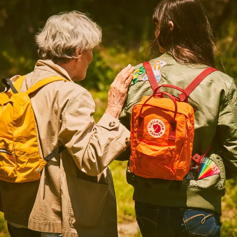 An elderly woman walks with her hand on the shoulder of a young girl wearing a backpack.