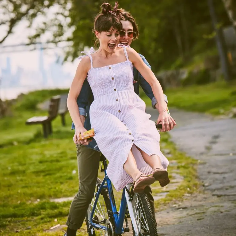 A woman in a white sundress rides on the handlebars of a man riding a bike.