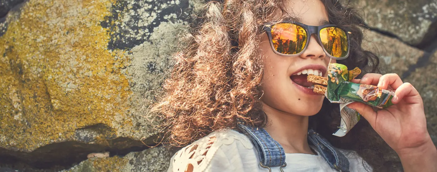 A curly haired girl wearing sunglasses, leaning against a rock wall and eating a Nature Valley Crunchy bar.