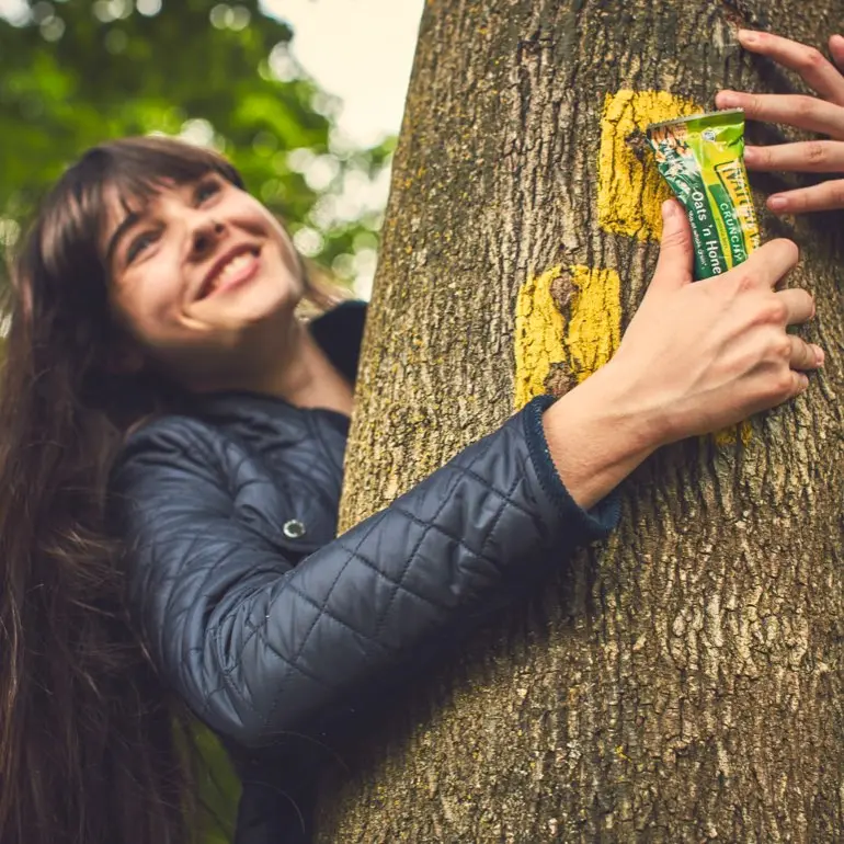 A smiling dark-haired woman hugs a tree while holding a Nature Valley bar.