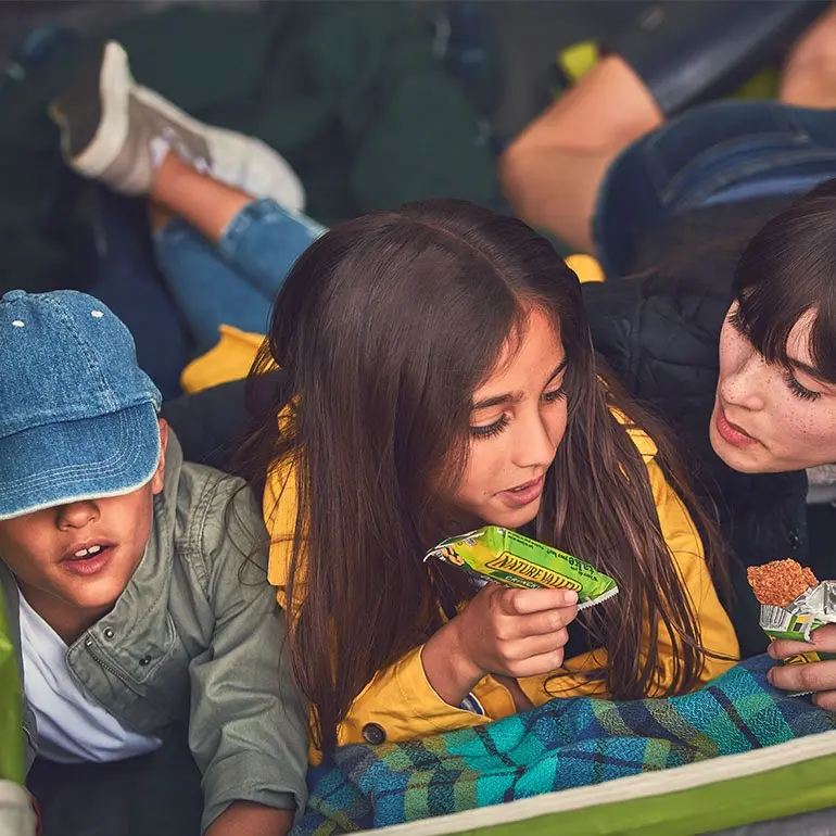 Three kids laying side by side on blankets in a tent eating Nature Valley granola bars.