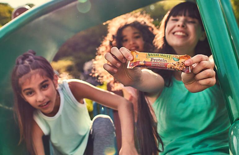 Three kids enjoying the outdoors whilst one holds a Nature Valley Peanut Butter, Dark Chocolate Protein bar.