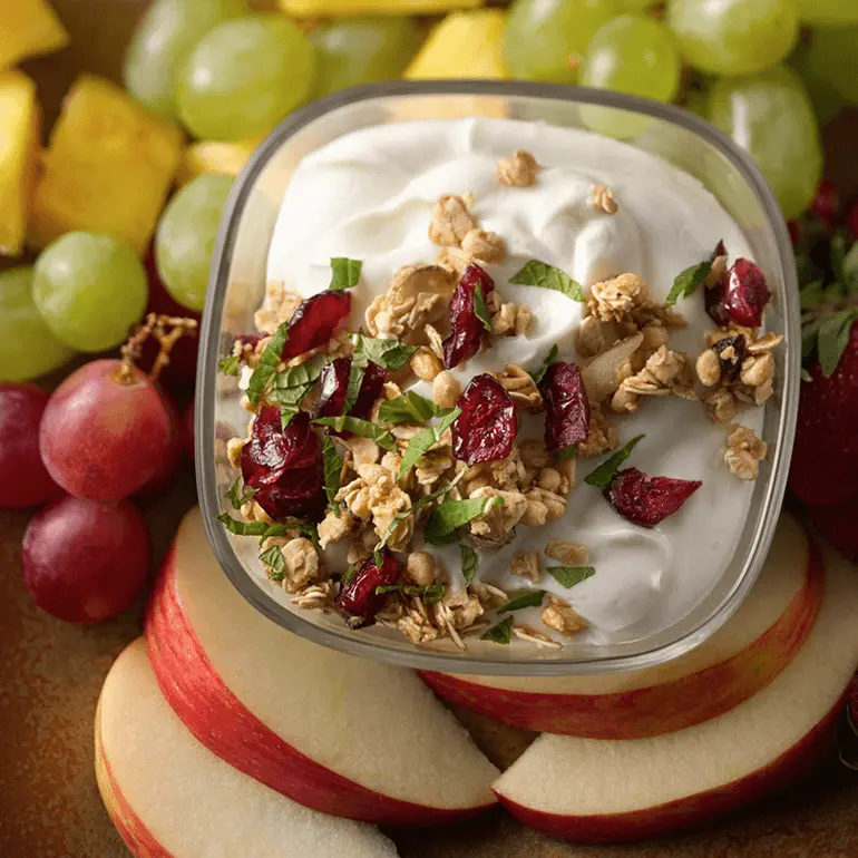 Greek yogurt dip topped with Nature Valley Cranberry Almond Protein Granola and chopped fresh mint leaves amongst apple slices and green grapes.