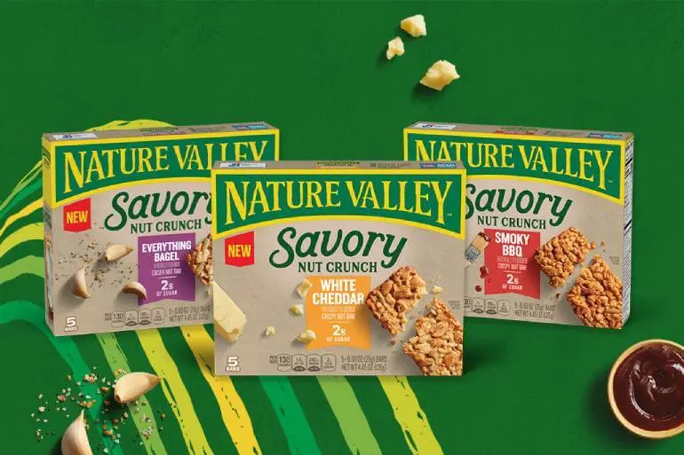 A family shot of three Nature Valley Savory Nut Crunch Bars, Everything Bagel, White Cheddar, and Savory BBQ on a green background with a yellow and green line pattern and a cup of BBQ sauce and garlic cloves.