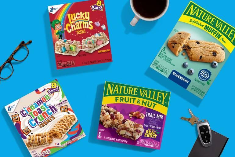 A collection of front facing General Mills products such as Cinnamon Toast Crunch Treats, Lucky Charms Treats, Nature Valley Soft-baked Muffin Bars and Nature Valley Fruit & Nut Trail Mix on a blue background with a pair of glasses, car keys, an apple and a cup of coffee.