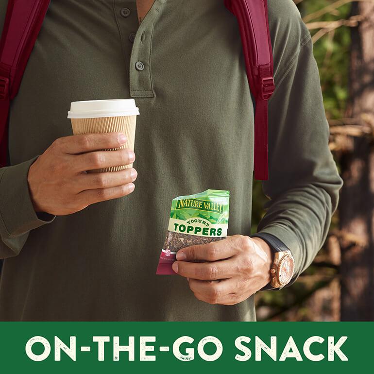 A close up of a man in a green top holding a coffee cup and a pouch of Nature Valley Yogurt Toppers Dark Chocolate Cherry flavor.