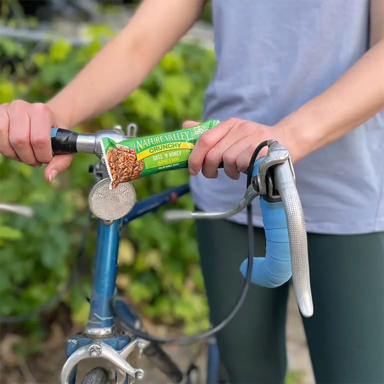 A person in a gray shirt holds a partially opened Nature Valley crunchy oats and honey granola bar while standing next to a blue bicycle. Green foliage is visible in the background.