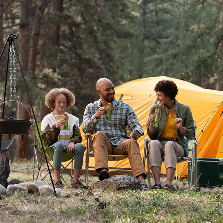 A family of three sits in front of a yellow tent in a forest, smiling and holding drinks. They are seated on camping chairs near a campfire area with a cast iron pot hanging above it. Trees are visible in the background.