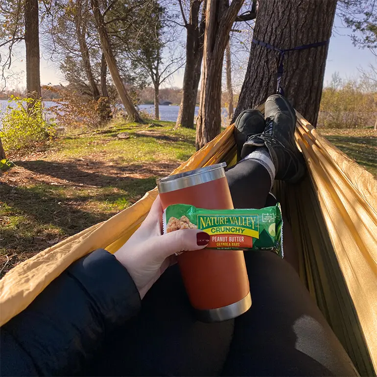 Person relaxing in a hammock by a lake, holding a cup and a Nature Valley crunchy peanut butter bar. Theyre wearing black pants and shoes, with trees and water visible in the background under a clear sky.