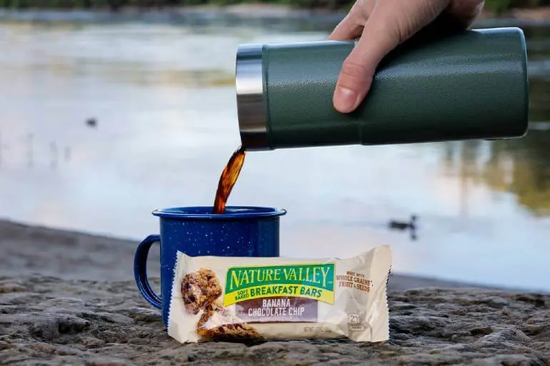 A hand pours coffee from a green thermos into a blue mug on a rock surface by a lake. In front of the mug is a Nature Valley banana chocolate chip breakfast bar. The background shows a calm lake and distant trees.