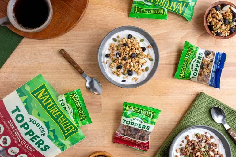 A breakfast scene featuring a bowl of yogurt topped with granola. Various Nature Valley granola packets are scattered around. A cup of coffee is on the side, and a green napkin with a spoon rests nearby on a wooden table.