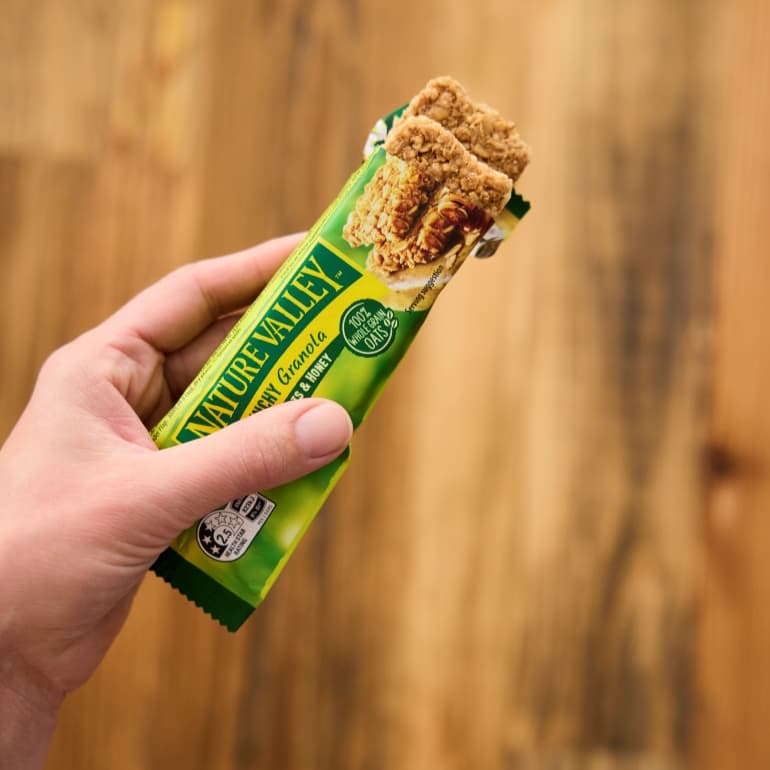 Hand holding an opened Nature Valley Crunchy Granola Oats & Honey bar wrapper, with two oat granola bar pieces visible at the top against a blurred wooden background.