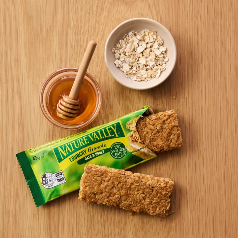 Nature Valley Crunchy Granola Oats & Honey bars displayed on a wooden surface, with one wrapped bar and two granola bar pieces alongside a small bowl of rolled oats and a dish of golden honey with a honey dipper, highlighting the product’s key ingredients.