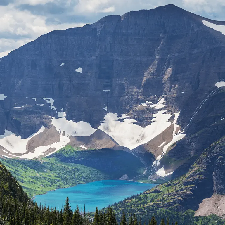 Lake in Glacier National Park, surrounded by grass, trees, and a snowy mountain.